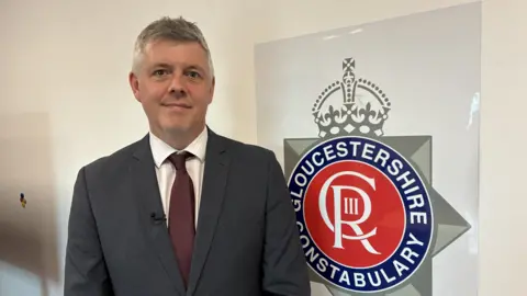 DCI Matt Phillips standing in front of a Gloucestershire Constabulary sign. He has short grey hair is wearing a dark-grey suit with purple tie.