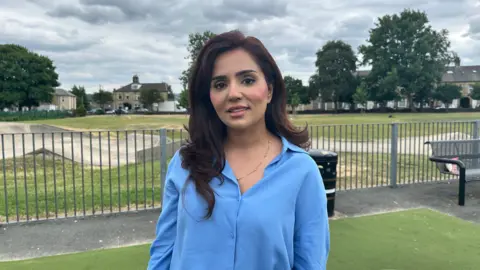 A woman with shoulder-length brown hair, wearing a light blue blouse. She is stood in a park, and behind her is a skating area, grass and trees.