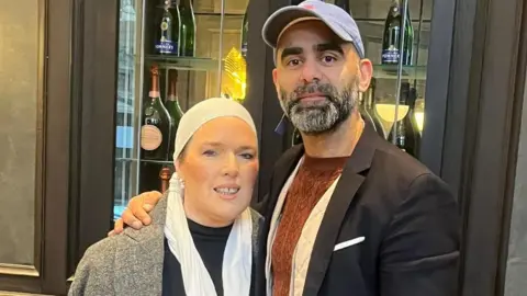 Eve Hawwa Iqbal-Khokhar, who wears a light head bandana and grey coat, poses next to her husband at a restaurant. Her husband wears a black jacket over a brown t-shirt and has a blue cap on. They both look at the camera.