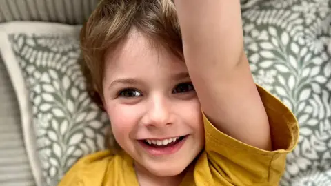 Axel, aged 5, sitting on a sofa. He is smiling and wearing a yellow T-shirt