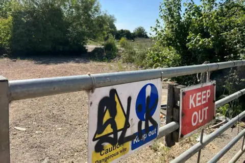 John Devine/BBC A farm gate with signs saying "keep out" and "caution - dangerous site". There is black graffiti on one of the signs. There is gravel, bushes and trees behind the gate in the background.