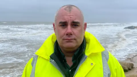 Edd Smith/BBC Daniel Candon is standing in front of the sea and is wearing a green shirt, a green jacket, and a yellow hi-vis jacket.