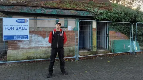 Simpa Carter is standing in front of the fenced-off toilet block. The one-storey brown-brick building has a triangle roof and green door and window panes. A FOR SALE sign is attached to the fence. Simpa is wearing black jeans and black hoodie with red sleeves, and a cap.
