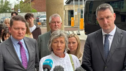 Shows a woman with grey hair and a white top standing in front of microphones. She if flanked by two men in suits and other people are in the background.