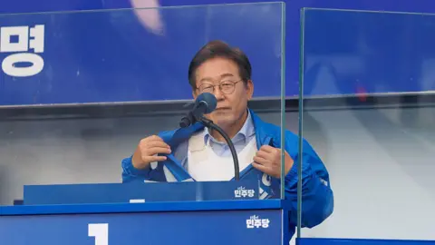 Getty Images Standing at a podium behind bulletproof glass, Lee Jae–myung pulls open a blue tracksuit jacket to show a bulletproof vest underneath