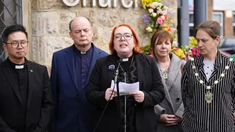 PA Media Reverend Catherine Hutton (third left) speaks to the media outside Epsom Methodist Church in Ashley Road, Surrey, following an alleged rape. She is with a group of people standing outside a church, with floral tributes visible on the wall behind them.
