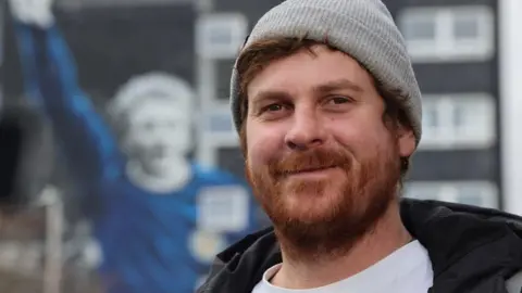 Man - artist Jerome Davenport - with beard, smiling at camera, with high-rise in background showing mural of footballer Denis Law.