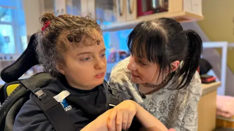 Amelie, who is strapped to a specialist device, has curly hair tied back and is holding her hands together as her mother looks on.