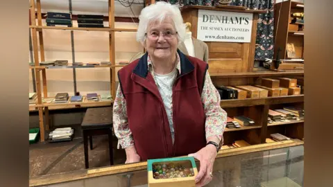 A woman with grey hair stands behind a glass display counter inside an auction room. The person is holding a shallow box filled with assorted marbles. Behind the counter are wooden shelves containing small boxes and display cases. A sign reading “DENHAM’S – The Sussex Auctioneers” is visible on the wall above.