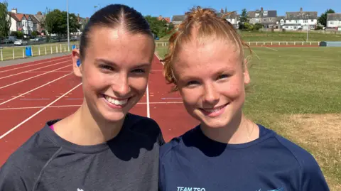Honor and Getta smiling into the camera while on a red running track. Honor is on the left and has brown hair which is tied back. She has a blue hearing aid in her ear and is wearing a grey t-shirt. Next to her, Gretta has strawberry blonde hair which is also tied back and is wearing a blue t-shirt. There is a patch of grass in the middle of the track and houses in the background.