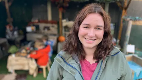 Georgina who has brown curly hair and is wearing a green coat and pink t-shirt is smiling at the camera. Behind her there are two children sitting a table outside busy playing.