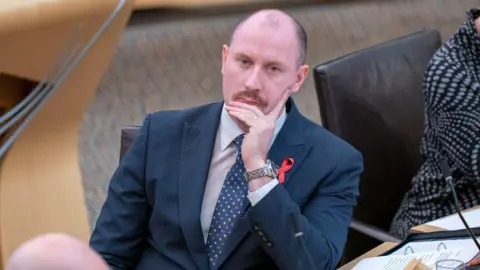 PA Media Neil Gray in the Holyrood chamber. He is resting on his chin in his hand and is looking thoughtful. He is dressed in a blue suit with tie, and has a moustache.