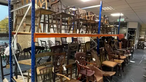 Wycombe Museum A large selection of old wooden chairs are seen piled on and near to racking shelves in a storage area