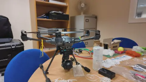 A modern drone sits on an office table. Surrounding it are lines of plastic-wrapped syringes, a tennis ball which was stuffed with drugs, bags of pills and some electronic cables.