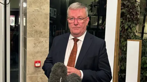 PA Mike Nesbitt stood in front of microphone wearing navy suit, white shirt and orange tie, in building lobby with plants, windows and stone walls in background