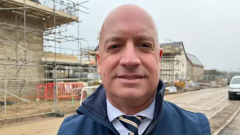 Mr Evans is stood amongst building work on the housing estate. The buildings have scaffolding on them and the road is largely unmade. He is wearing a blue zip up jacket, a pale shirt and a striped tie. 