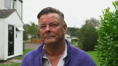 A man stands on green space near a house, wearing a purple fleece and a white shirt. His hair is slicked to the side and he is looking slightly off camera.