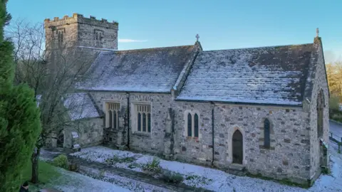 Stephen Nichols Exterior of the 15th-century stone church of St John the Baptist at Spetisbury following a dusting of snow. The church walls are primarily flint with large brown-looking stones laid at intervals creating a random decorative effect. It has a pitched, tiled roof and a square crenelated tower. The doors and windows are stone gothic arches and the glasswork is lead came.