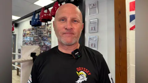 Zac Sherratt/BBC Ross Getty, head coach at Odyssey Boxing Club in Stanwell, wearing a black t-shirt with the club logo, and is bald. He is standing in a boxing ring. 