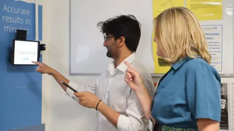 CUH Jamie Mistry is standing with Louise Allen in front of an eye test screen in a hospital. He has dark hair and a beard and is wearing a light-coloured shirt. He is holding a mobile phone in his left hand. Louise Allen is standing behind him. She has fair hair and is wearing a blue blouse