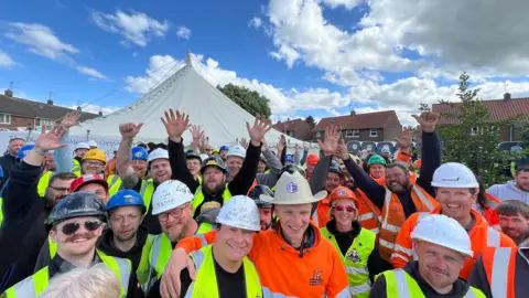 Amanda White/BBC A group of construction workers wearing hard hats and high-visibility vests. They're gathered outdoors under a blue sky with white clouds. They are smiling and raising their hands in celebration. A large white tent and residential houses are in the background.