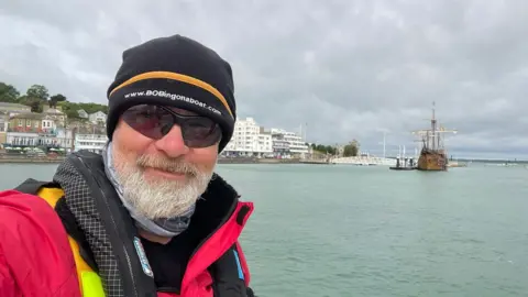 Clipper Ventures Bob Brown, wearing a life jacket, in front of the sea, with a coastal town in the background
