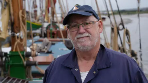 Tim Kenney is dressed in a navy sailor's jacket, wearing a navy baseball cap, and standing on the deck of Pudge the Thames sailing barge