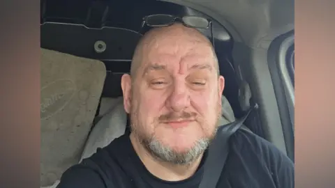 Mike Beale Mike Beale is sitting in his van and smiling slightly at the camera. He has very short grey hair, almost bald. He has a short beard and moustache. His wearing a black t-shirt and his glasses are perched on top of his head.