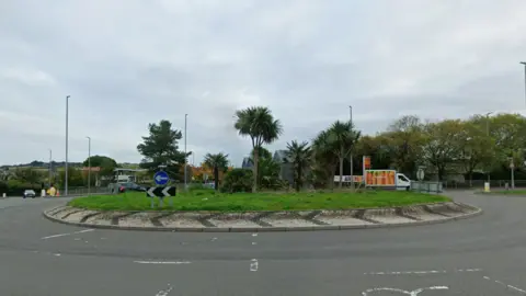 The roundabout on the A390 on a cloudy day. The islands has palm trees and grass as well as road traffic signage. There are two cars and a van on the opposite side of it and the lanes in the foreground are empty. 