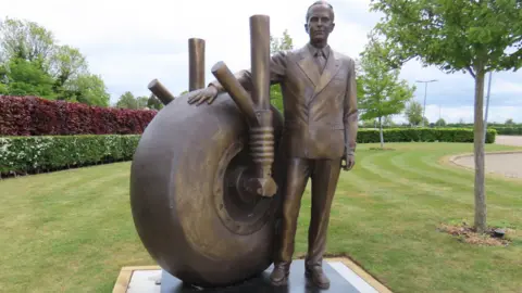 The statue is bronze in colour and Sir George wears a suit and tie. He has his hand on the top of the wheel. The statue is on a plinth in the manicured gardens of the IBCC.