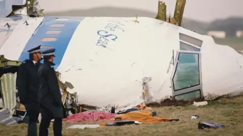 Getty Images The right hand side of the nose cone of Pan Am Flight 103 is embedded into the ground in a field. Metal struts jut from the wreckage. Debris including blankets covers the ground. Two police men in large coats and caps are standing looking at the debris.