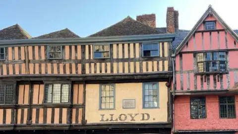 John Bassett A row of tudor style buildings. They are orange, yellow and red with black panels on them. The yellow building in the middle has "Lloyd" written on it in capital letters