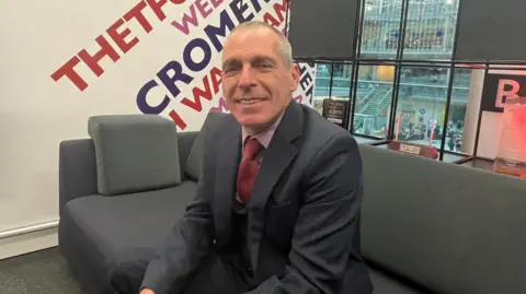 Nic Rigby/BBC Peter Dutch is sitting on a grey sofa in a BBC studio. He is smiling at the camera and is wearing a dark suit, pink shirt and red tie.
