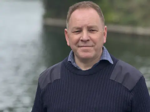 Councilor Richard Coe is standing by the River Thames near the bridge linking Windsor to Eton. Wearing a blue jumper over a checked blue shirt you ca see the water behind him but nothing else is in the shot.