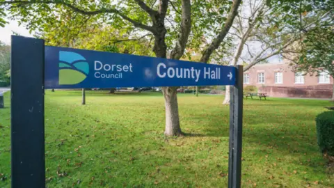 A Dorset Council sign pointing towards County Hall in Dorchester, with County Hall in the distance. 