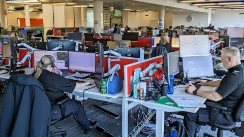 Staff working at their desks in Durham Police's control room. A blonde-haired woman has her back to the camera as she looks at her computer screens. A grey-haired man is sitting to her right. There are dozens of desks across the room.