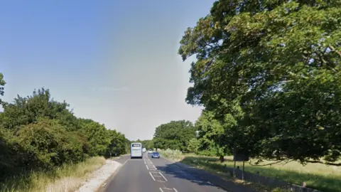 Google A single carriageway main road in the countryside, with chevrons down the middle. There is a layby on the left side and trees either side.