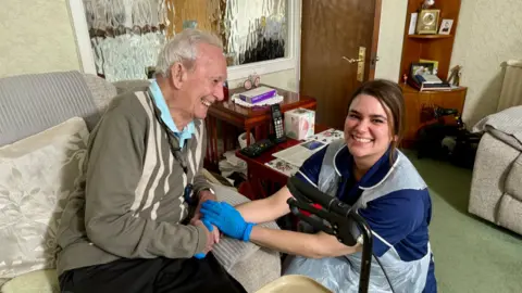 STEVE HUBBARD/BBC Joana Curvelo crouches next to her 104-year-old client John. He is sitting on a grey velour sofa and is wearing a khaki and cream cardigan. In the background is a small wood dresser with a few shelves on it and some family photographs. Jonana has long brown hair, dark brown eyes and is looking at the camera. She is wearing dark blue scrubs with a white trim and a plastic apron and blue gloves, John is smiling at her and is hold her hands