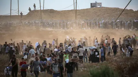 Getty Images A crowd of Palestinians waiting in a long line at one of the aid sites in Gaza as dust rises up all around them, while in the background a handful of armed figures overlook the scene from the top of a mound of earth.