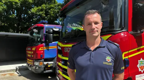 A man with short cropped hair stands in front of two red fire engines. He is wearing a dark blue shirt with a Humberside Fire and Rescue Service logo on it.