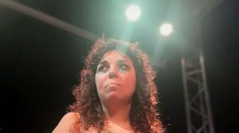 Bee Wilkes Uma garota com cabelos pretos cacheados parece triste sob as luzes do palco