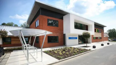 EPUT The front of the St Aubyn Centre which is a red brick building with a cycle rack outside. There are rainbow coloured windows to the side and it has a few plants in a bed to the front.