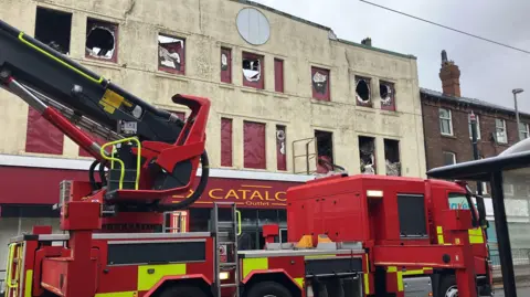 A close up of a fire engine in front of the empty former three-storey Ex Catalogue Outlet building on Lord Street, Fleetwood, taken the morning after a fire. Most of its windows are damaged by the fire.