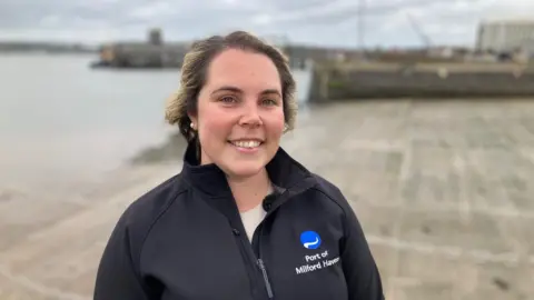 Image of Claire Lawrence in a zip up coat in front of Pembroke Harbour. 