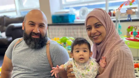 Nausheena Nackwa and her family. From left to right a brown skinned man with a bald head. He's a black and grey beard, and is wearing a brown back pack, over a grey t-shirt. He's sitting down next to his six month old daughter, who has black hair and is wearing a flowery bib. On the far right is a woman wearing a pink headscarf. 
