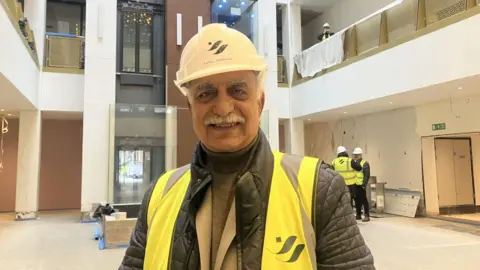 Mahboob Hussain, wearing a white hard hat, and a high visibility jacket. He is standing in one of the restaurant rooms smiling.