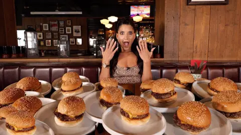 A woman sits in a restaurant with several plates with burgers on the table in front of her. She has straight dark hair and she is hold her hands up with a mock shocked expression.