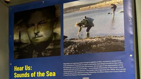 MNH The display, with photographs of the project. One black and white image shows a the face of a man wearing a shirt and tie, who has a moustache, next to a lighthouse. Another image shows two women standing in the sea with recording equipment. Yellow writing reads Hear Us: Sounds of the Sea.