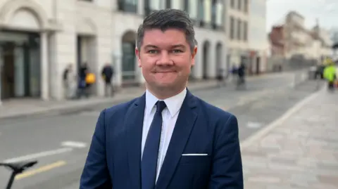 BBC A photo of Jersey politician, Sam Mézec. He is standing in a street wearing a blue suit with a blue tie and a white shirt. He is looking straight at the camera. The background is partially blurred but you can see a road, pavement and some people walking behind him. 