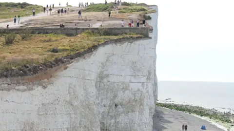 A white chalk cliff, with people stood close to the edge taking photos. Below, there is a walkway with people walking on it, and the sea in the background.
